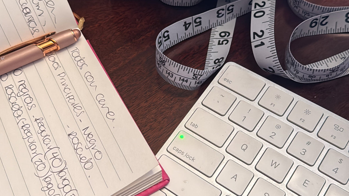 A black fabric scrunchie with a white measuring tape wrapped around it, styled on a wooden desk next to a pink notebook, pen, and white keyboard.
