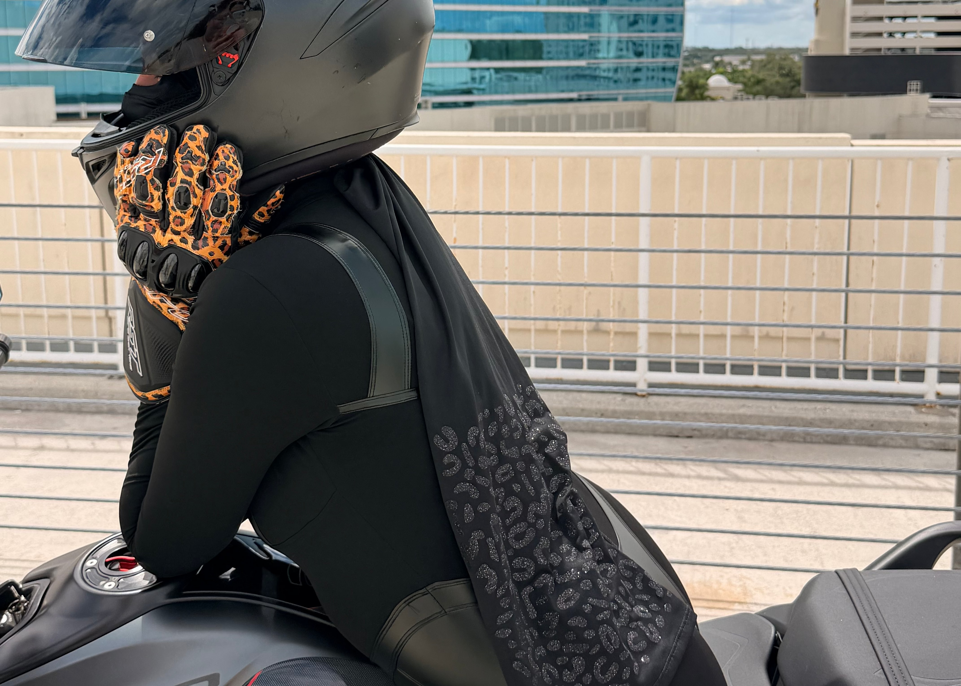 A person wearing a matte black motorcycle helmet, black riding jacket, and black-and-orange patterned gloves sits on a dark gray motorcycle outdoors with a modern blue glass building in the background.