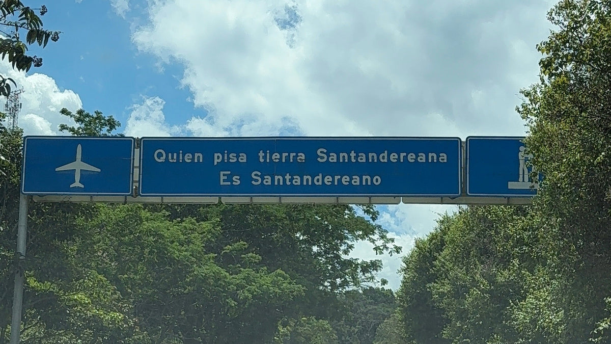A view from inside a vehicle on a road under a blue sky with clouds, featuring a large blue overhead highway sign with white text and a motorcycle driving ahead.